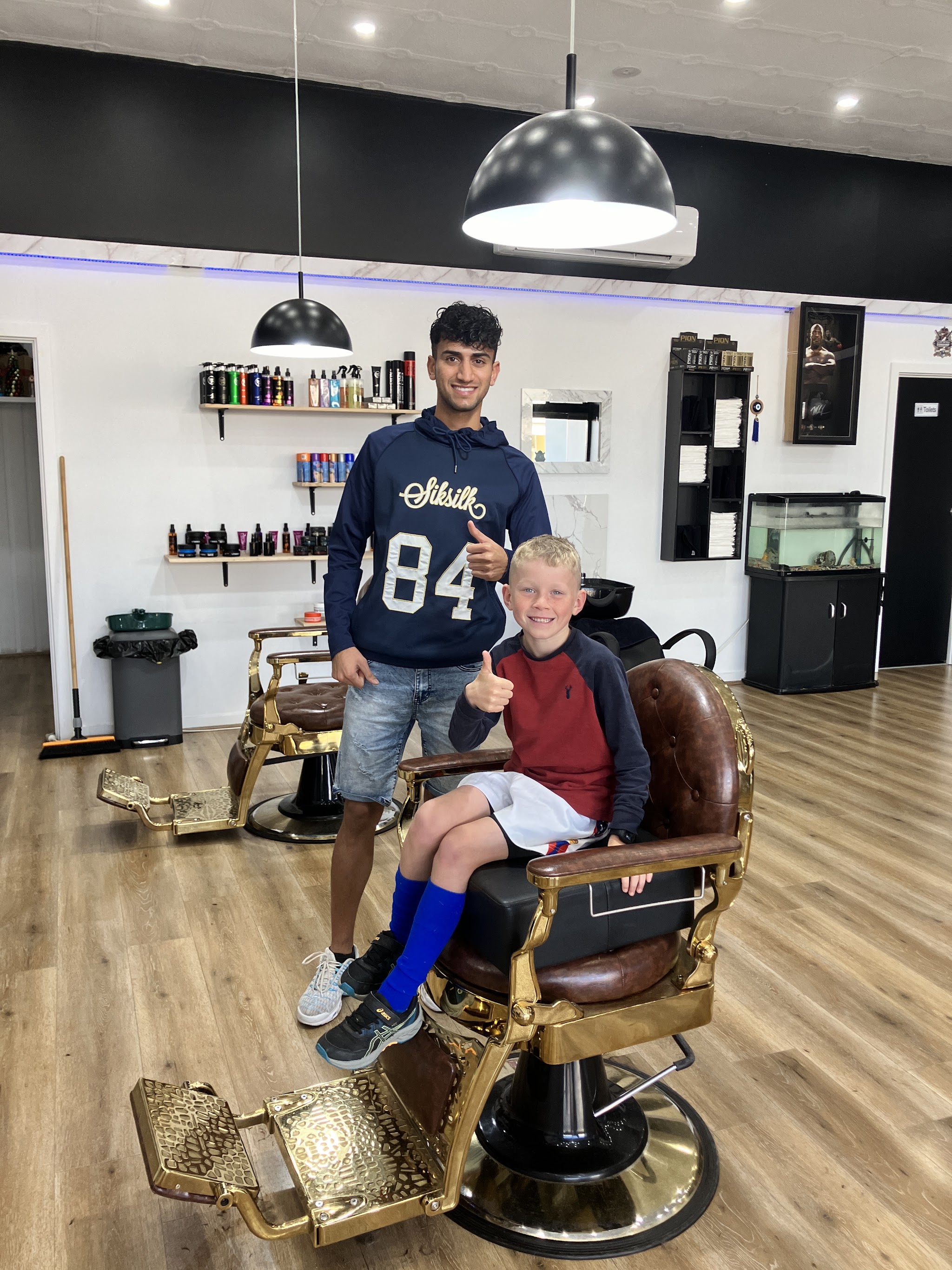 A barber and a young customer giving thumbs up from a vintage gold barber chair